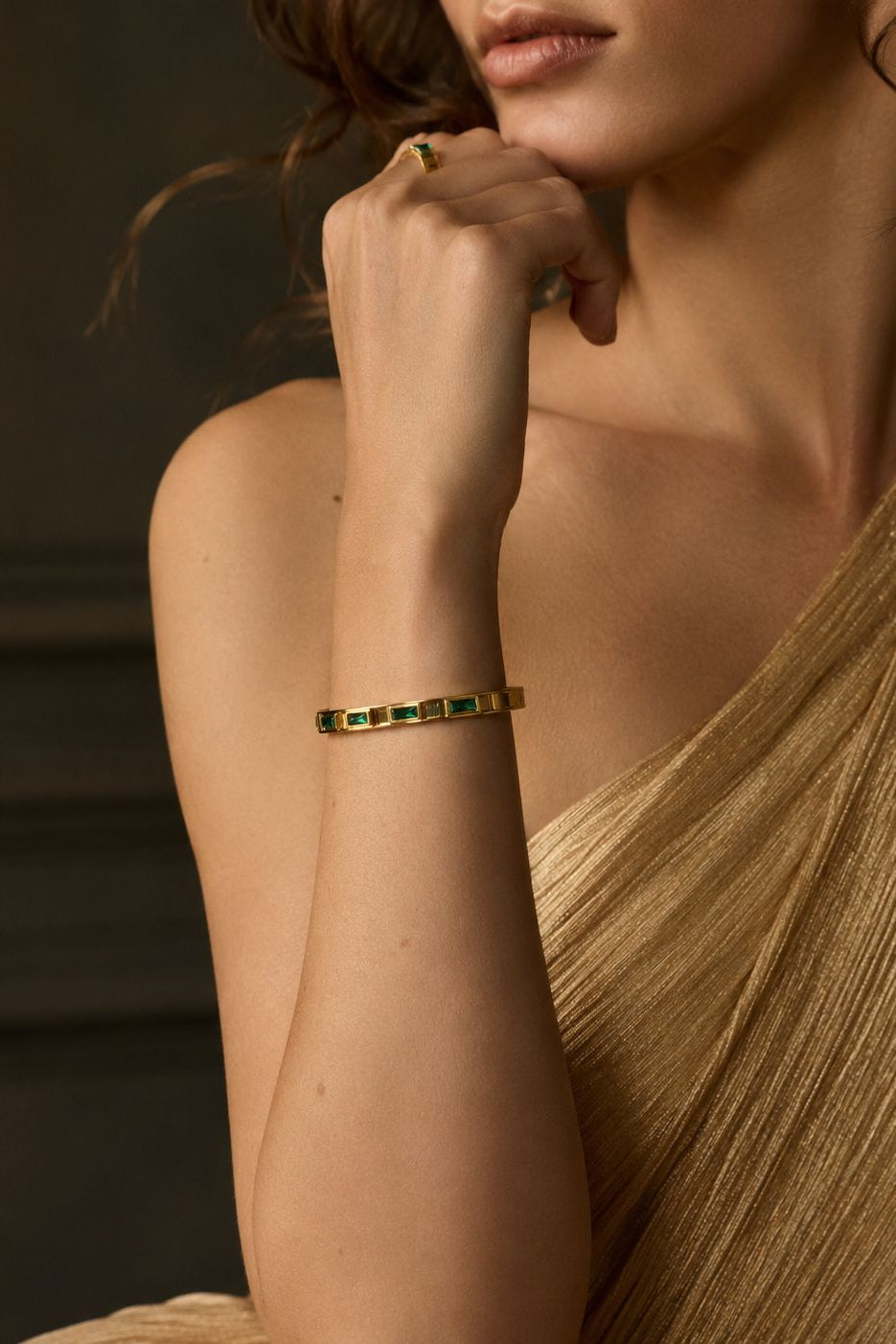 Woman wearing a gold bracelet with green gemstones against a dark background